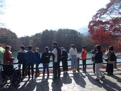 紅葉が美しい湖のほとりで、児童たちが並んで景色を眺めている様子の写真