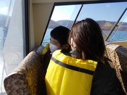 ライフジャケットを着た児童が、観光船の窓越しに湖の景色を眺めている様子の写真