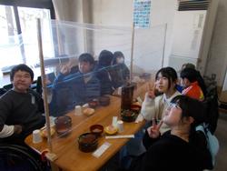 児童たちがアクリル板で仕切られたテーブルを囲み、食事前に楽しそうにピースサインをしている様子の写真