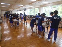 オープンスペースに複数置かれた机に分かれて生徒たちが炊き出し訓練を行っている様子の写真