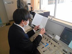 放送室でマイクに向かって原稿を読んでいる男子生徒を後ろから写した写真