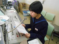 放送室でマイクに向かって原稿を読んでいるショートカットの女子生徒を横から写した写真