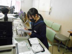 放送室でマイクに向かって原稿を読んでいる髪の毛を一つに束ねた女子生徒を横から写した写真