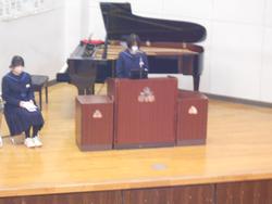 グランドピアノが置かれた舞台で演台に立った女子生徒がマイクに向かって話をし、その左後ろの椅子にもう一人の女子生徒が座っている写真