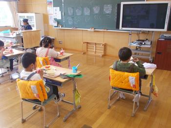 小学校の教室で、子どもたちが間隔をあけて座り、黒板の方向を向きながら給食を食べている様子の写真