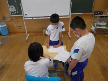 ジャージ姿の児童3名が机の周りで一人が座り、一人が鉛筆で記入をしている様子を写した写真