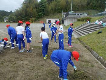 赤白帽子を被りジャージ姿の児童達が校庭で草をかき集めている写真