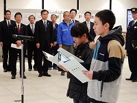 多くの大人が見守る中、マイクの前に立つ2人の子供が、真剣な表情でクリップボードに挟んだ書類を読み上げている様子の写真