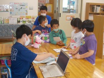 一人の男子児童がパソコンの前に座り、机の反対側に5名の児童が並んでいる図書室内での写真