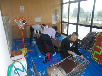 ブルーシートを敷いた室内で長机やパイプ椅子の準備をしている3名の男性の写真