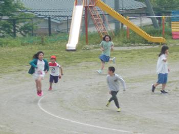 5名の児童が滑り台などの遊具が置かれた校庭を走り回っている写真