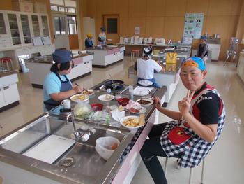 児童と調理台に向かい合わせに座り、料理を食べている右側のエプロン姿の女性が笑顔でピースをしている写真