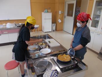 エプロン姿の男の子と女の子のペアで、女の子が盛り付けの準備をし、男の子が料理が入ったフライパンの横に立っている様子を写した調理実習の写真