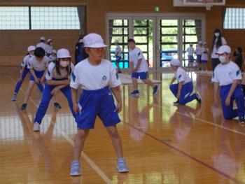 体操服を着た4人の児童が、体育館に引かれた白線をまたいで立ち、その横に測定をする生徒たちが並んでいるスポーツテストでの写真