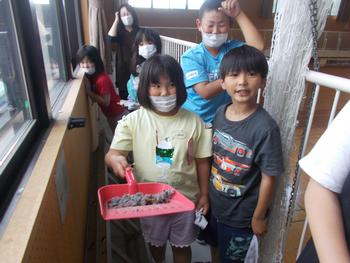 掃除をした児童たちが窓の横の通路に集まり、手前に立った女子児童が塵取りに乗せたたくさんのホコリをカメラに見せている写真