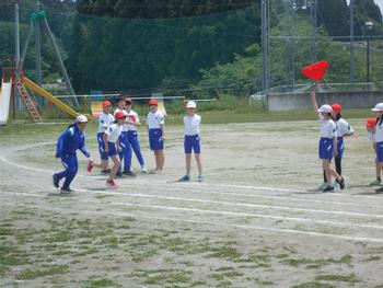 複数の児童が見学する中、スタート位置で赤い旗を持ってスタートの合図を送る児童と、トラックに並んだ2人の女子児童が走り出した様子を写した写真