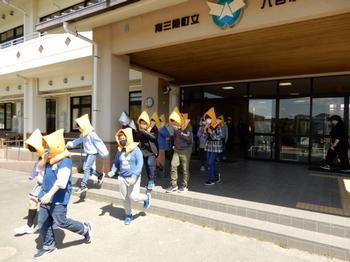 頭に防災ずきんをかぶった児童たちが列をなして正面玄関前より外へ避難しているところを写した写真