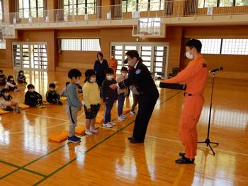 体育館で小学生の子どもたちが数人、横一列に並んで前を向いて立ち黒っぽい制服姿の男性が1人ずつ子どもに手を差し出し、賞状や記念品のようなものを手渡しをしており、そばにはオレンジ色の制服を着た消防隊員らしき人物が立っている写真
