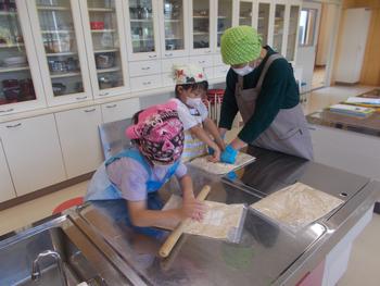 エプロン姿の2名の女子児童が綿棒や手を使って、講師の人の手伝いをもらいながら、袋に入れた大豆をつぶしている写真