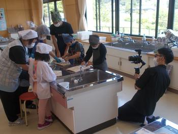 カメラを持った男性が横にしゃがんで撮影をする中、調理台で数名の児童と講師の方が味噌づくりを行っている様子の写真