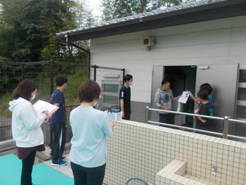 プール横の建物の前に数名の先生が集まり、メモをとりながらプールの機械の操作の説明を聞いている写真