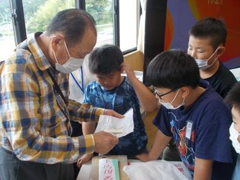 男性が白い紙を持ち、周りに男子児童4名が集まってその手元を覗き込んでいる写真