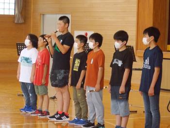 7名の児童が1列に並び、左から3番目の男子児童がリコーダーを演奏し、6名が合唱をしている写真