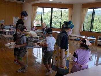 お盆をもった数名の児童が順番に部屋の中央の配膳係から給食を受け取っている様子の写真