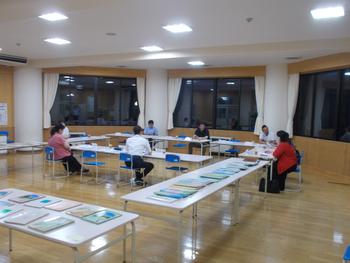 教室の窓際にロ型に長机が配置され数名の人たちが座り話し合いをし、教室手前の長机に資料が並べられている写真