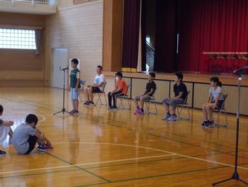 5名の児童が舞台前のパイプ椅子に座り、一人の男の子がマイクの前に立っている壮行式での写真