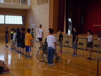 舞台の前の椅子の前に立つ6名の児童とその前に並んで立っている児童達の写真