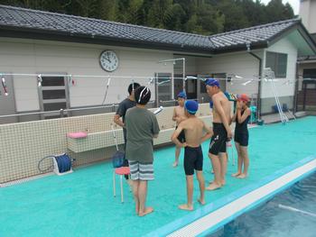学校の屋外プールのプールサイドで先生と子どもたちが輪になって立って先生の話を聞いている写真