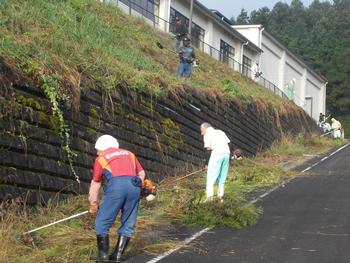 学校の周りの壁の草をビーバーで刈り取っている男性2名の写真