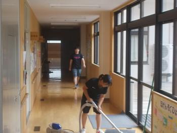 ろうかの奥を歩いている人と入り口のマットに掃除機をかけている関係者の写真