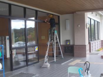 脚立にのりながら玄関入り口の扉の上部の掃除をしている関係者の写真