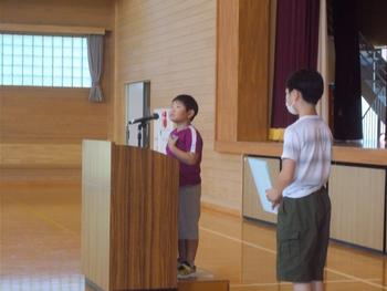 体育館の演台で一人の男の子がマイクを使い話をしている横で一人の男の子が立っている写真