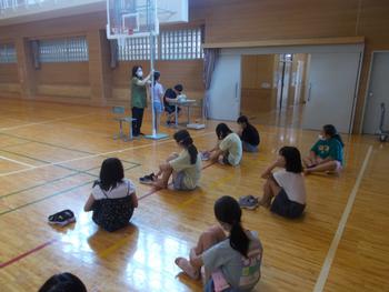 一人の女の子が体育館で身長測定を行い、他の女の子たちは床に座り待っている様子を写した写真