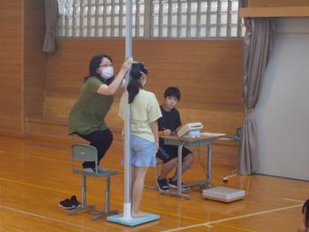 女性が椅子に乗りながら女の子の身長を測定し、机に座っているいる人が記録をしている様子の写真