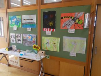 壁に7枚の絵と2枚の習字が展示され設置された長机のうえにもいろいろな作品が展示されている写真