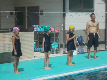 3名の女の子がプールのスタートラインに立ち、横に男性教諭が立っている校内水泳記録会での写真