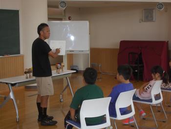 数人の子供が1番前で椅子に座り、目の前の男性の読み聞かせを聞いている写真