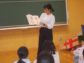 黒板の前に立ちながら絵本を子供たちに見せて読み聞かせをしている女性の写真