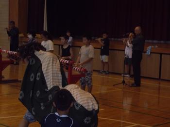 薄暗い体育館の中舞台の前で並んで笛を吹く生徒と指導する先生の写真