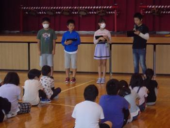 舞台前の4名の児童が並んで立ち、1人の男の子がマイクで話をしているのを床に座って聞いている生徒たちを後方から写した写真