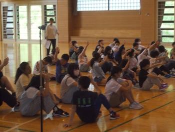 体育館の床に座っている生徒数名が手をあげている様子を写した写真