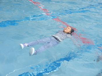 靴を履き洋服を着たまま1人の児童が上を向きプールの中で浮かんでいる写真