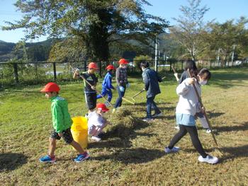 低学年から高学年の子までが帽子を被りほうきやレーキを使って草を集めている写真