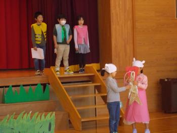 舞台に3名の児童が並んで立ち、階段下で2人女の子が劇をしている写真