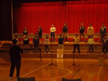 舞台の上に1列、その下に1列に並んだ子供たちが発表をしている写真