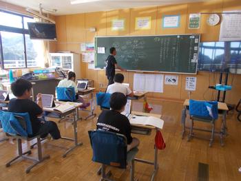 男性が黒板に字を書き、児童たちがタブレットを置いた机に座り授業を受けている写真